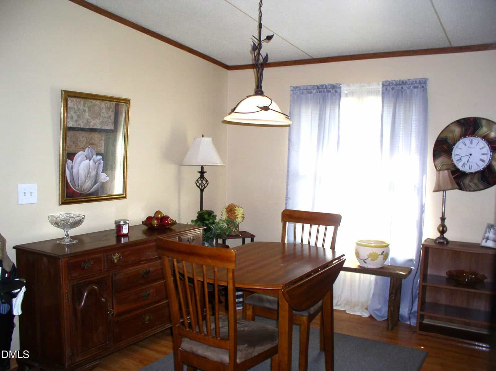Dining room with a wooden table and chairs, sideboard with decorative items, framed flower art, and a floor lamp. Light streams through sheer curtains by a window with a large wall clock and small table.