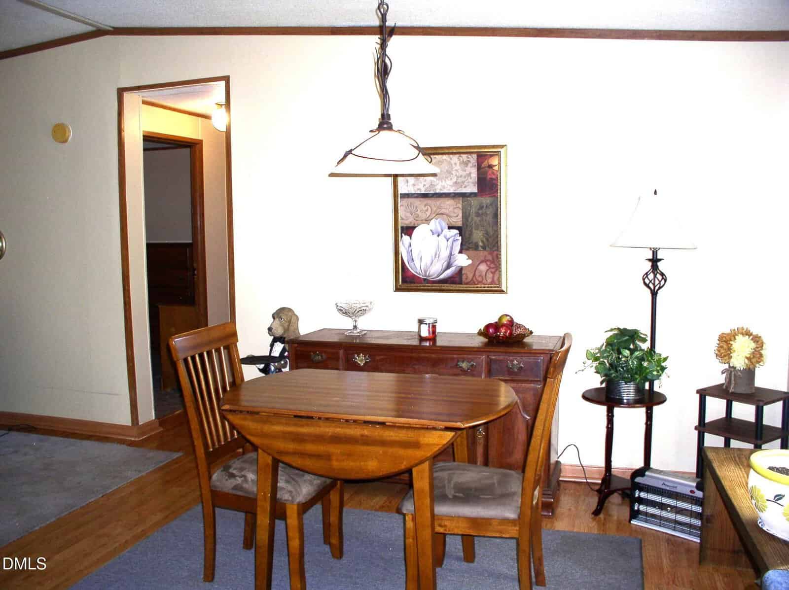 Dining area with a small wooden table and two chairs, a sideboard with decor items, a framed floral artwork, a floor lamp, and a potted plant on a stand, all set on a wood and carpet floor.
