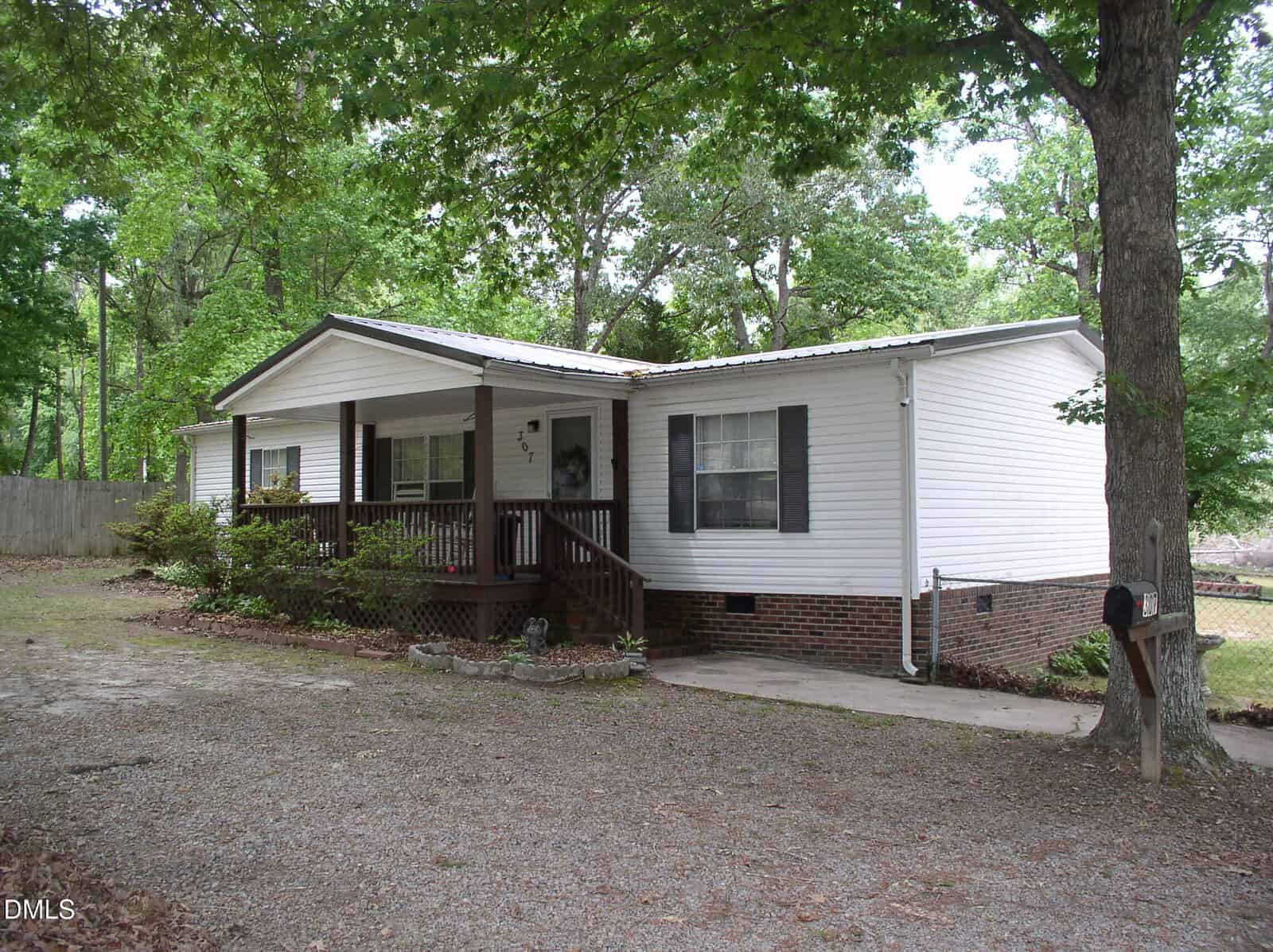 A white manufactured home with a brick foundation, covered front porch, and wooden railing. The house is surrounded by trees and has a gravel driveway in front.