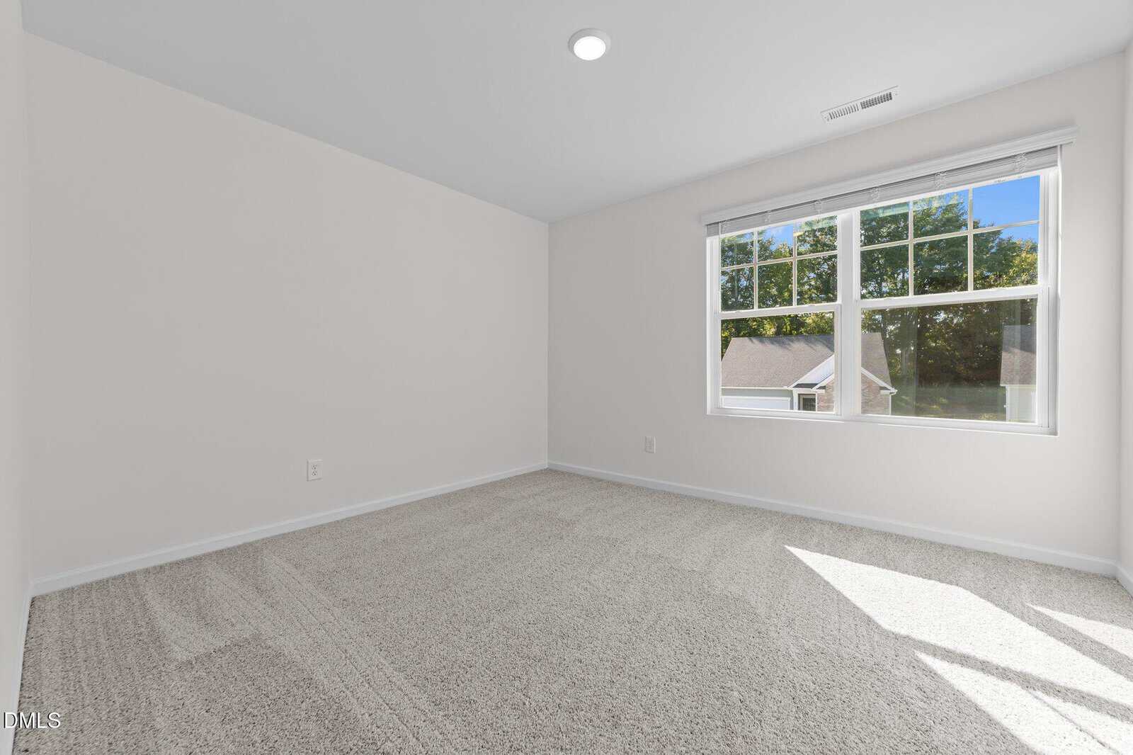 A bright, empty room with light-colored carpet, white walls, and a large window letting in natural light. The window offers a view of trees and rooftops outside.
