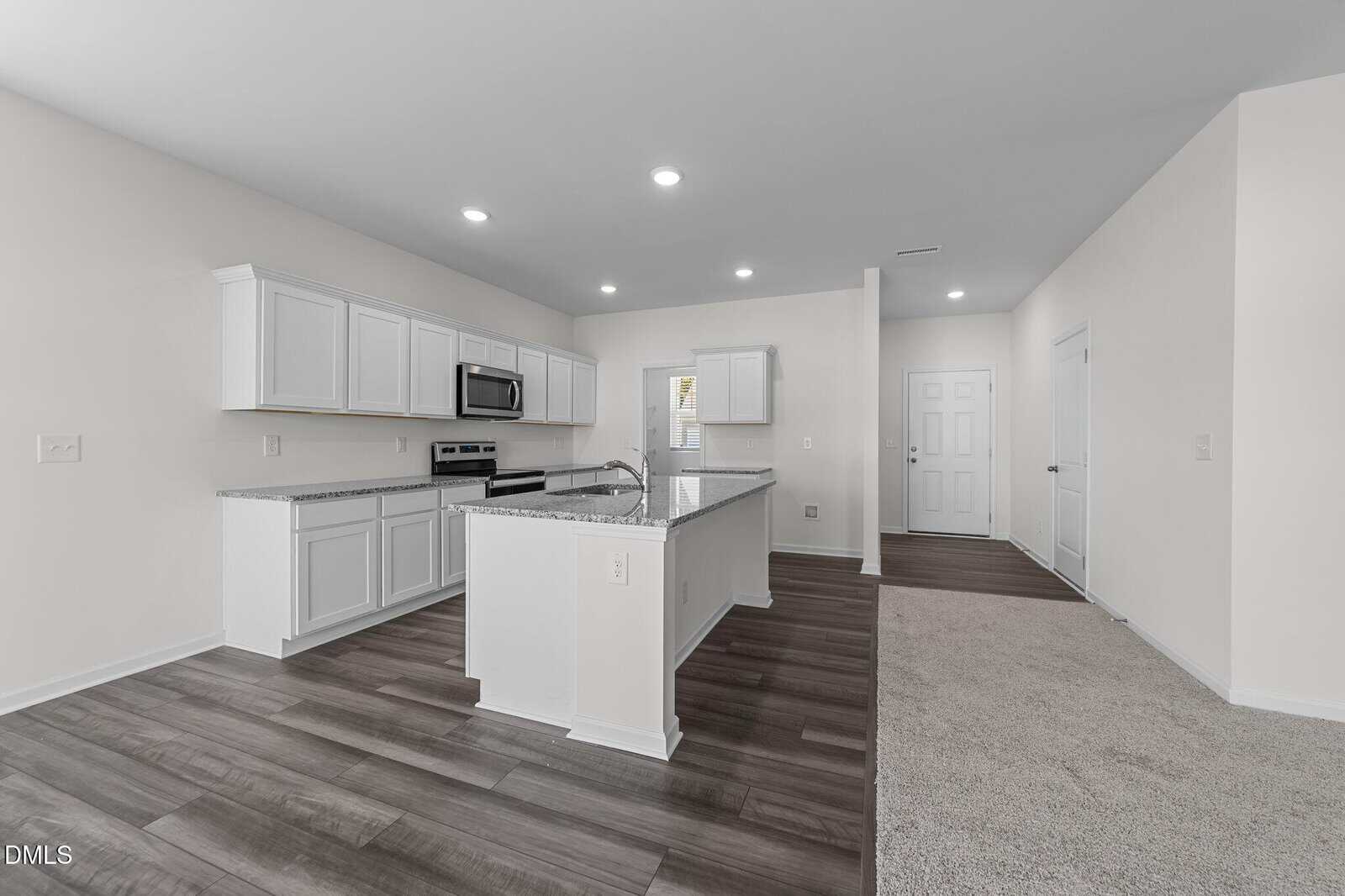 Modern kitchen with white cabinets, stainless steel appliances, and granite countertops, open to a living area with carpet and wood-style flooring, and a white entry door in the background.