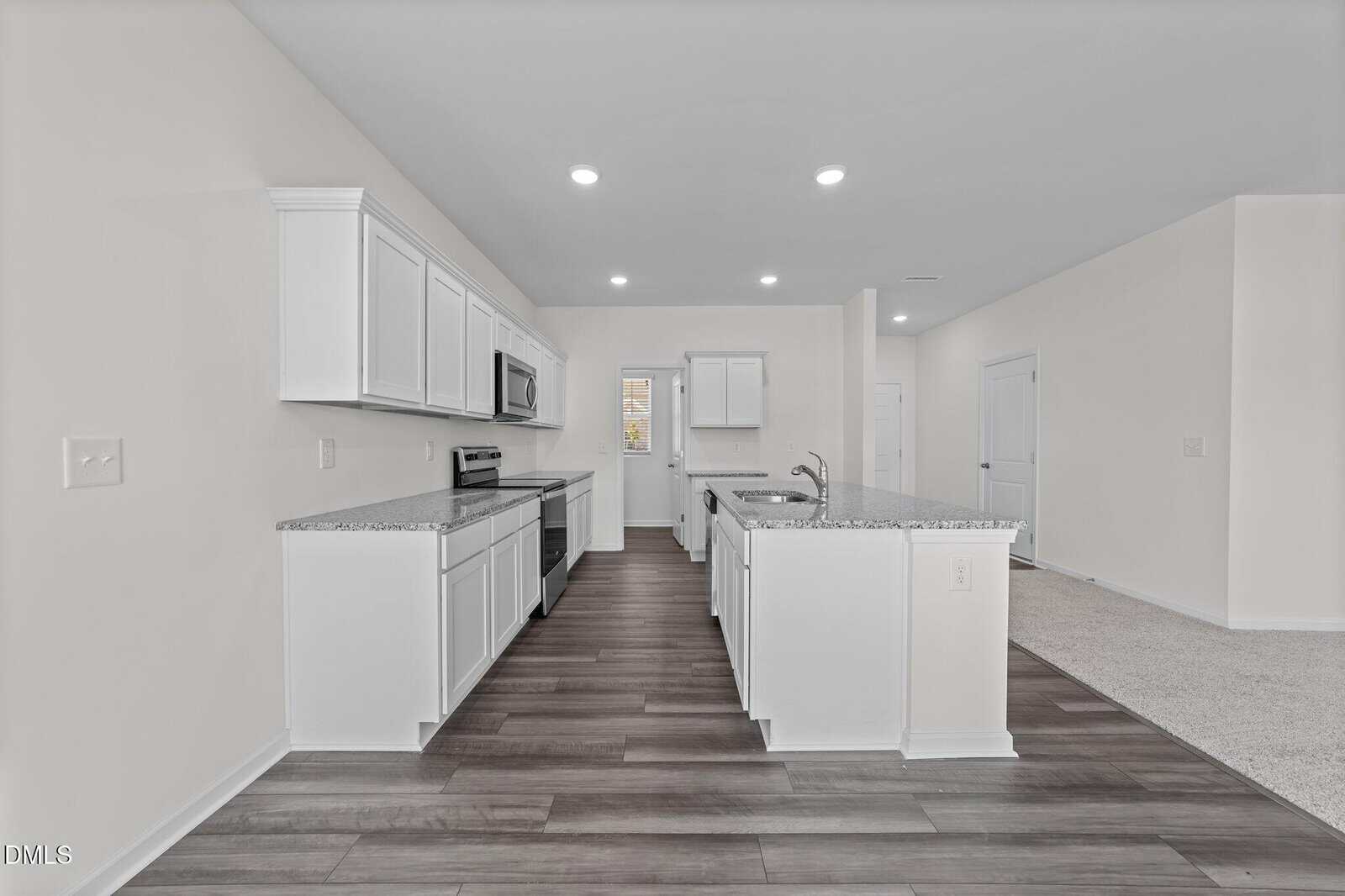 Modern kitchen with white cabinets, stainless steel appliances, granite countertops, and dark wood flooring. The area is well-lit with recessed ceiling lights and opens into a carpeted living space.