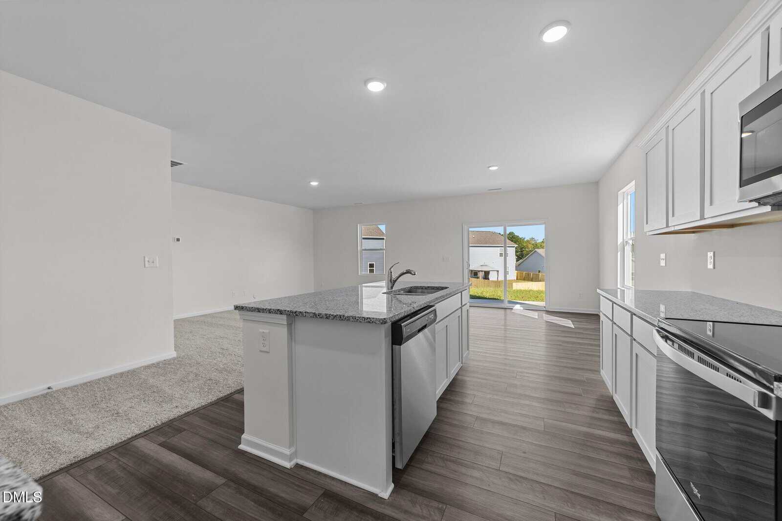 Modern kitchen with white cabinets, granite island, and stainless steel appliances opens to a bright living area with carpet and large windows, leading to a backyard visible through sliding glass doors.