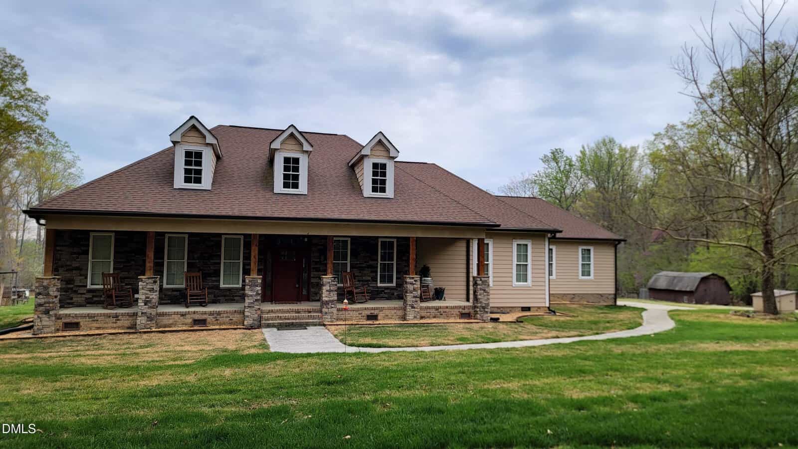 A large house with stone and beige siding, a brown roof, three dormer windows, and a covered front porch with chairs, set on a spacious lawn with a curved walkway and surrounded by trees.