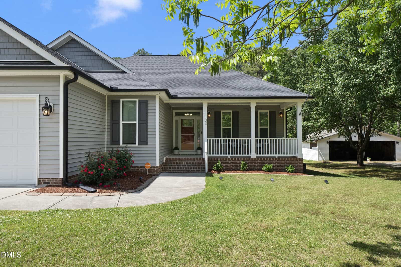 A light gray house with white trim, a covered front porch, and a manicured lawn. There are red flowers by the walkway, and a detached white garage is visible in the background among trees.