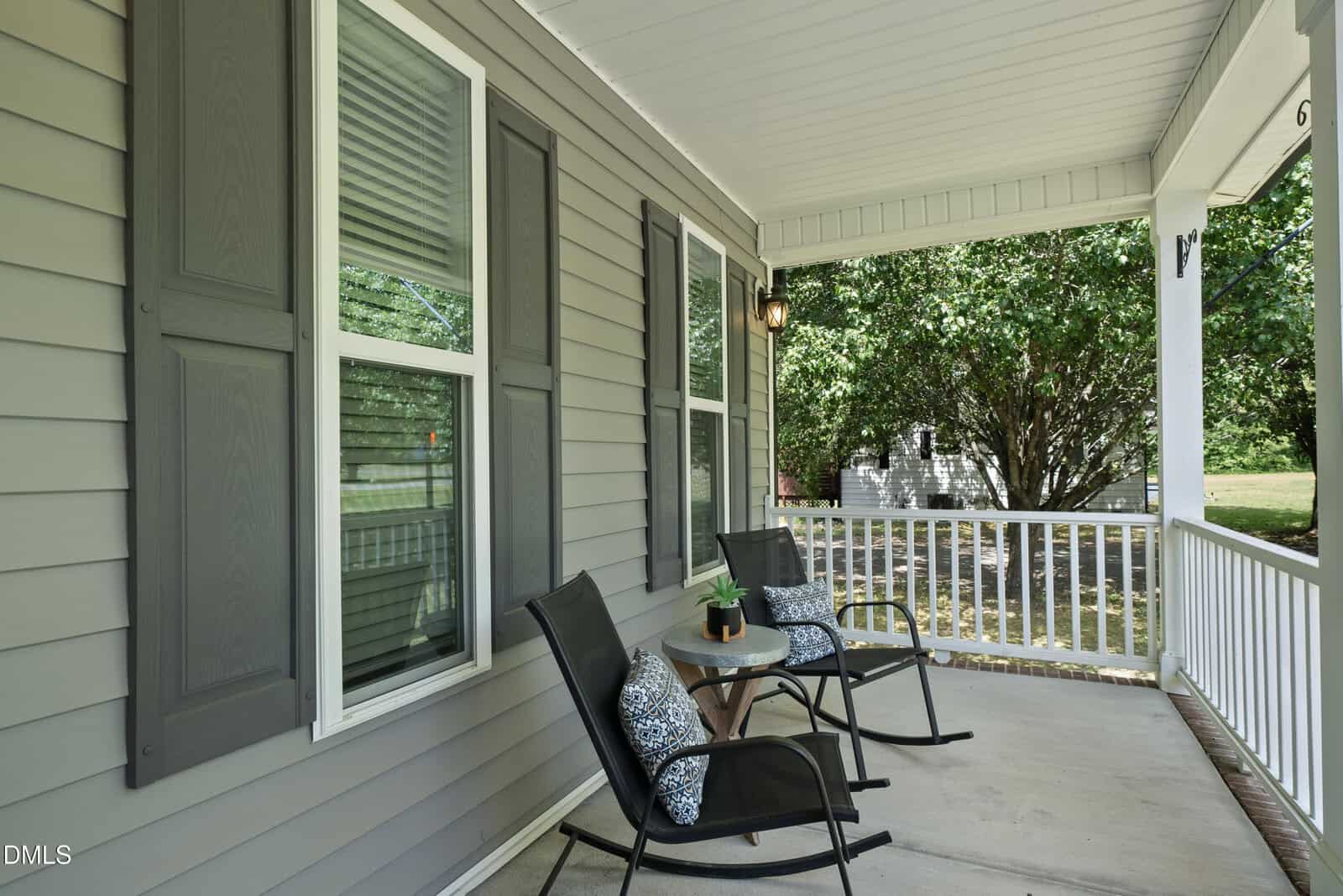 A cozy front porch with two black chairs and patterned pillows, a small side table with a plant, gray siding, white trim, and a view of green trees and a lawn beyond the white railing.