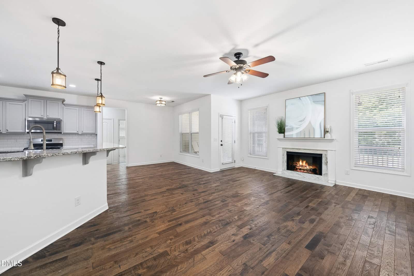 Open-concept living area with hardwood floors, a fireplace, two tall windows, and a ceiling fan. Kitchen with pendant lights and granite counters is visible on the left. White walls and natural light create a bright space.