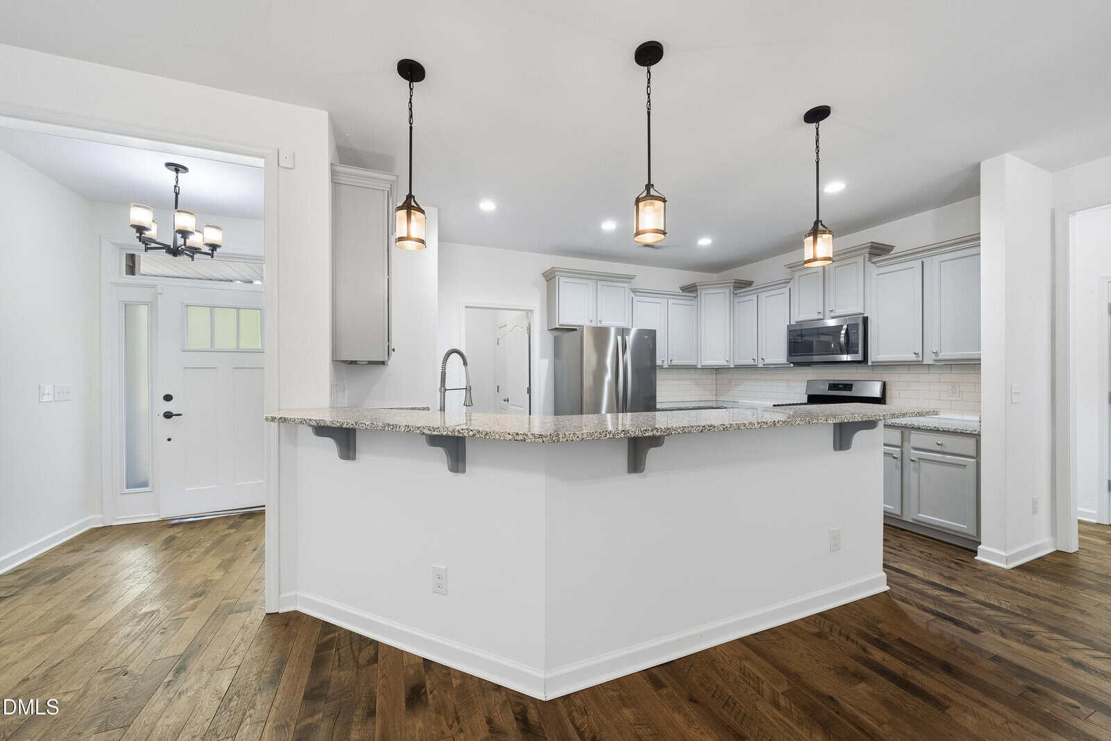 Modern kitchen with white cabinets, stainless steel appliances, granite countertops, pendant lights, and hardwood floors. An island with a sink faces an entryway with a front door and chandelier.