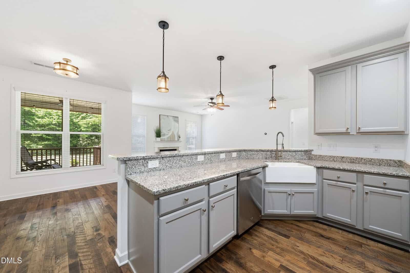 Modern kitchen with gray cabinets, granite countertops, farmhouse sink, pendant lighting, and stainless steel appliances. Open layout connects to living area with hardwood floors and large window showing a green backyard.
