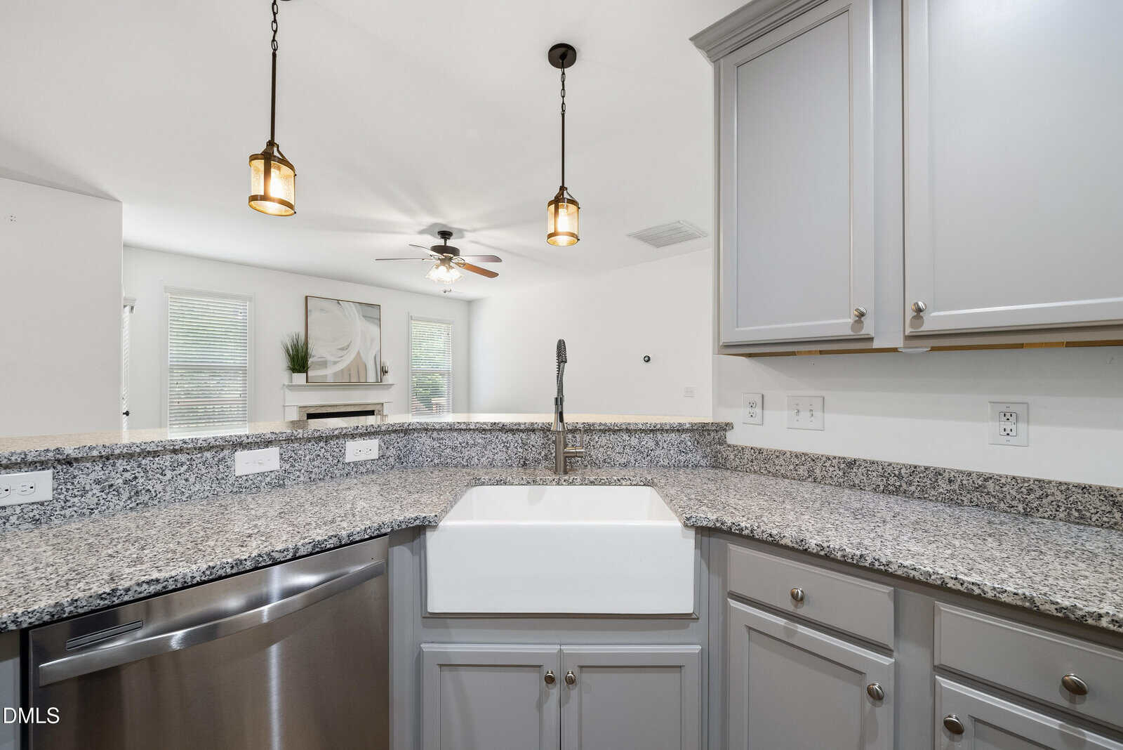 Modern kitchen with gray cabinets, granite countertops, a large white farmhouse sink, stainless steel dishwasher, pendant lights, and an open view to a living area with a ceiling fan, windows, and a fireplace.