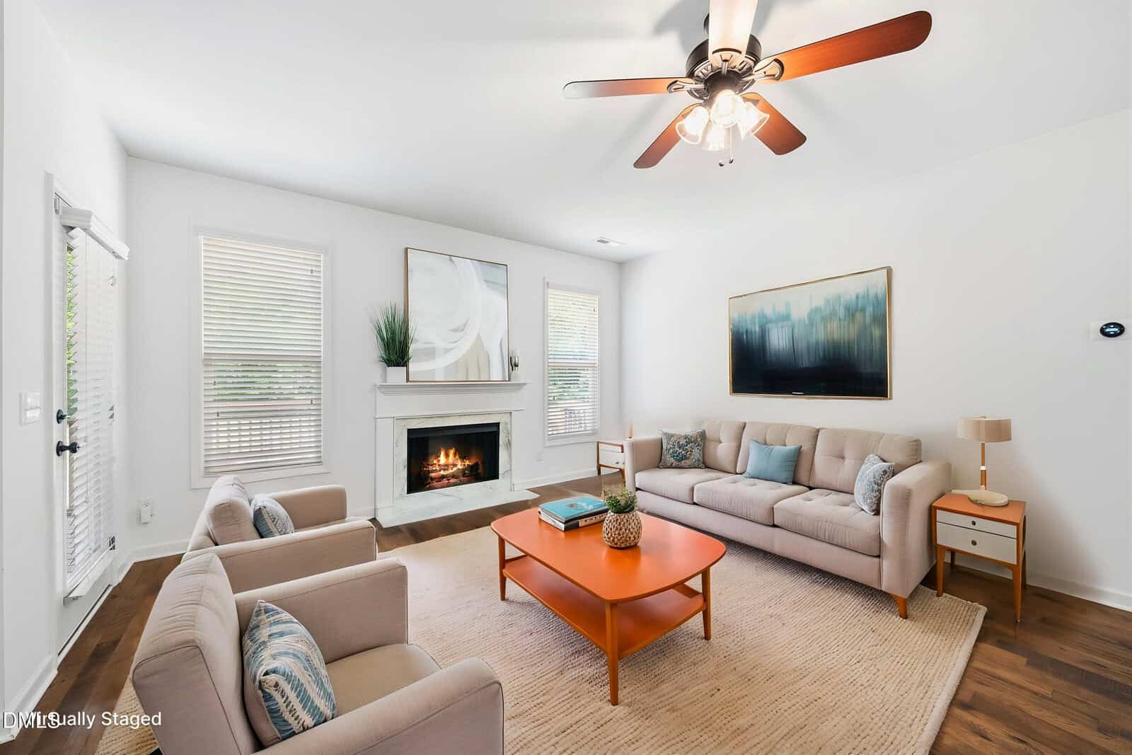 Bright living room with beige sofas and armchairs, a wooden coffee table, a fireplace with artwork above, large windows with blinds, and a ceiling fan with lights. Modern, minimal decor and cozy atmosphere.