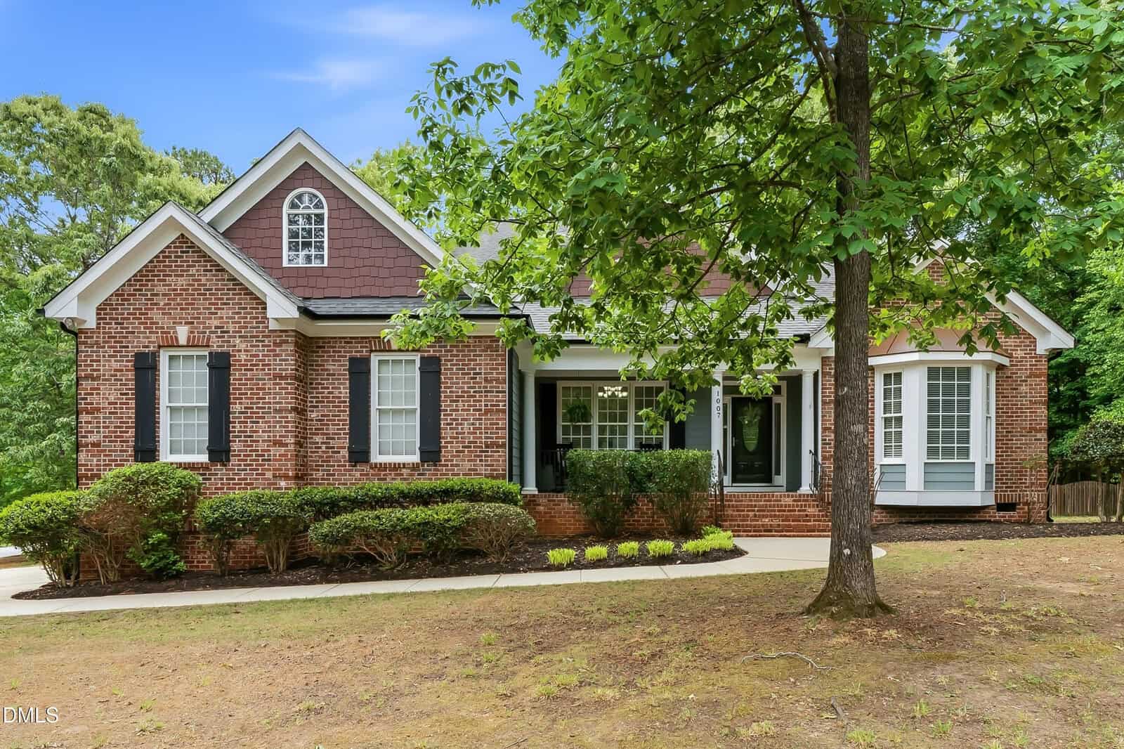 A single-story brick house with black shutters, white trim, and a gabled roof, surrounded by shrubs and a large leafy tree in the front yard. The house has a front porch and a bay window.