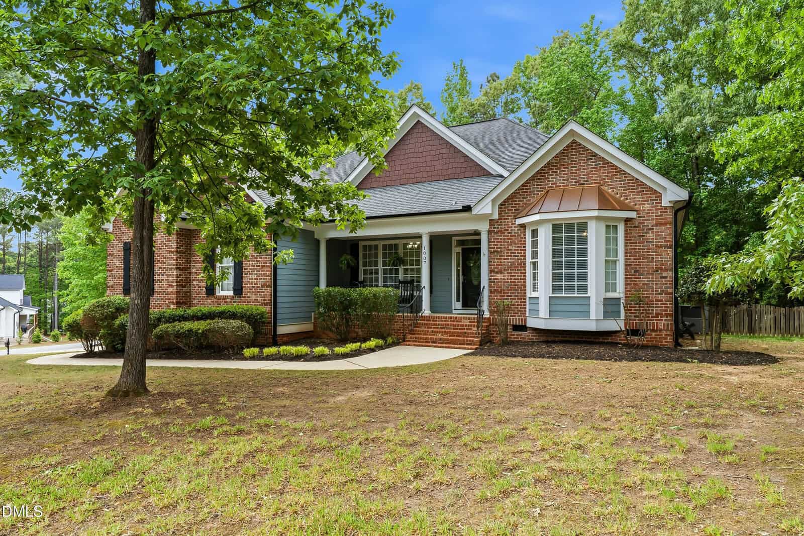 Single-story brick house with gray siding, a gabled roof, and a covered front porch. Large windows and a manicured lawn with trees and shrubs are in front; a clear blue sky is in the background.