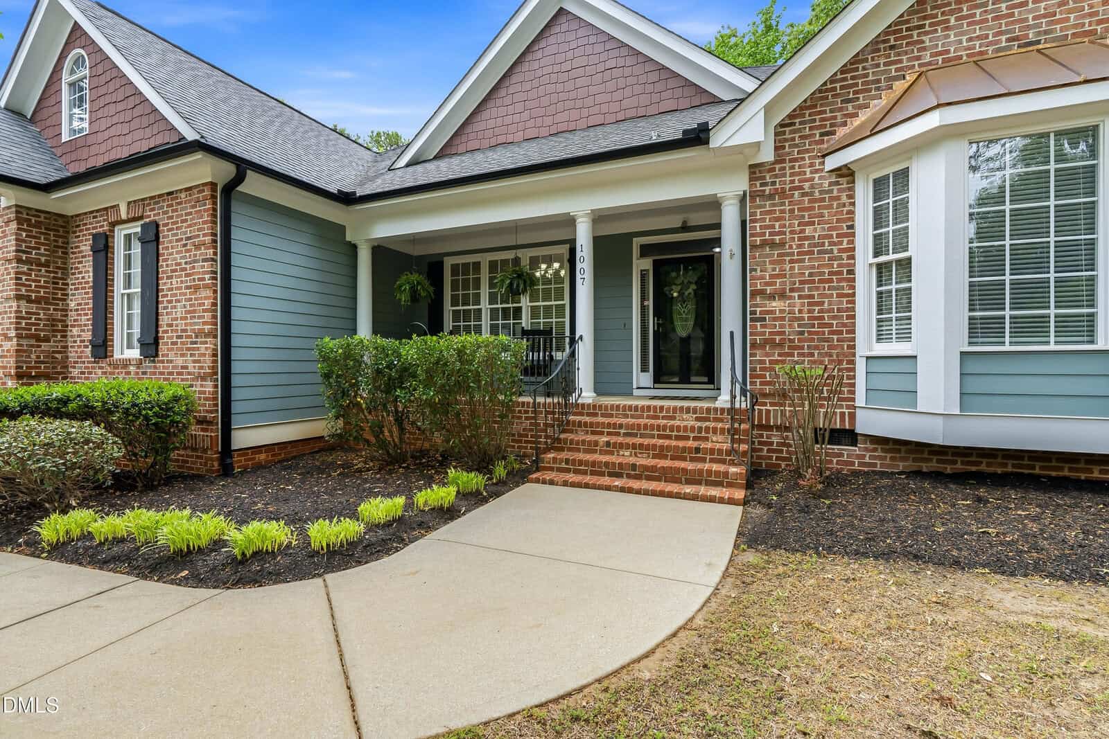 A front view of a house with a brick and blue siding exterior, white trim, a covered porch with steps, shrubs, and a curved walkway leading to the entrance.