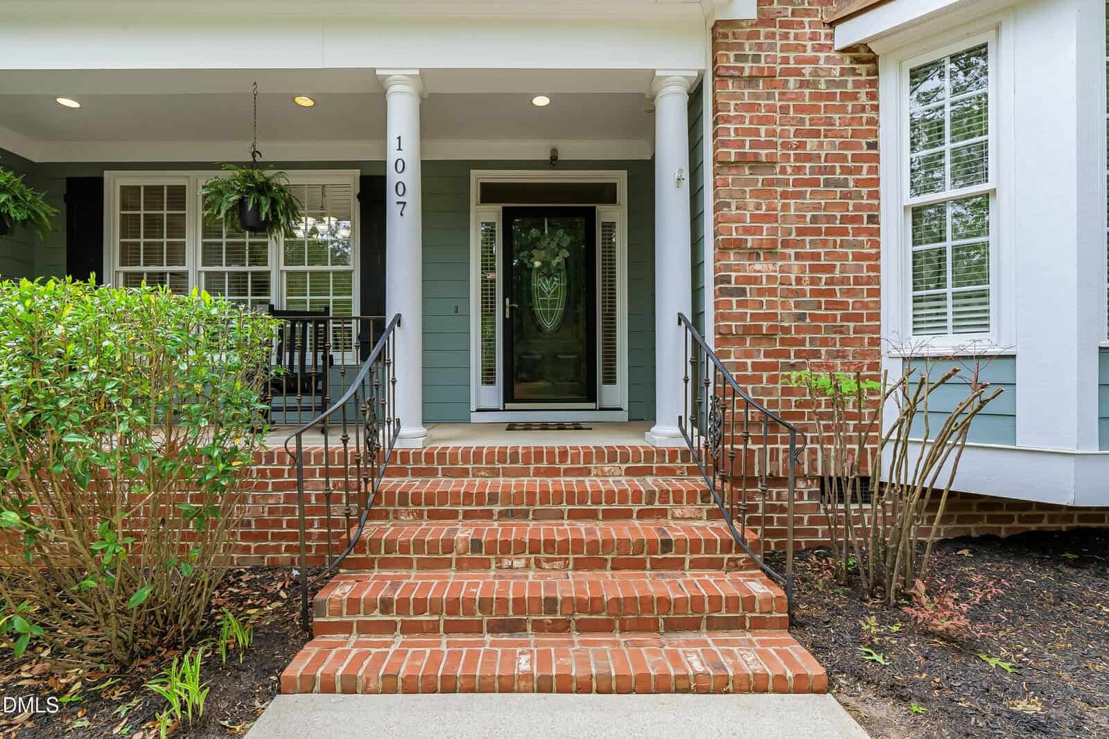 Front entrance of a house with red brick steps, black metal railings, two white columns, a glass door, green shrubs, and a hanging plant. The exterior walls are blue with white trim and some exposed brick.