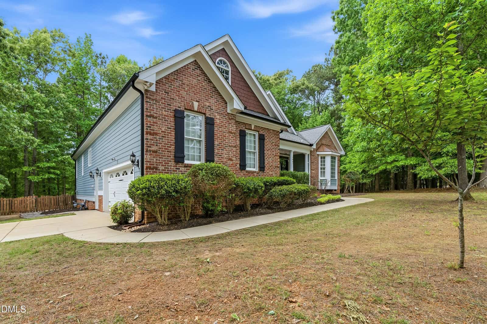 A brick and siding house with black shutters, a two-car garage, front shrubbery, and a curved driveway, surrounded by trees and a grassy yard under a blue sky.