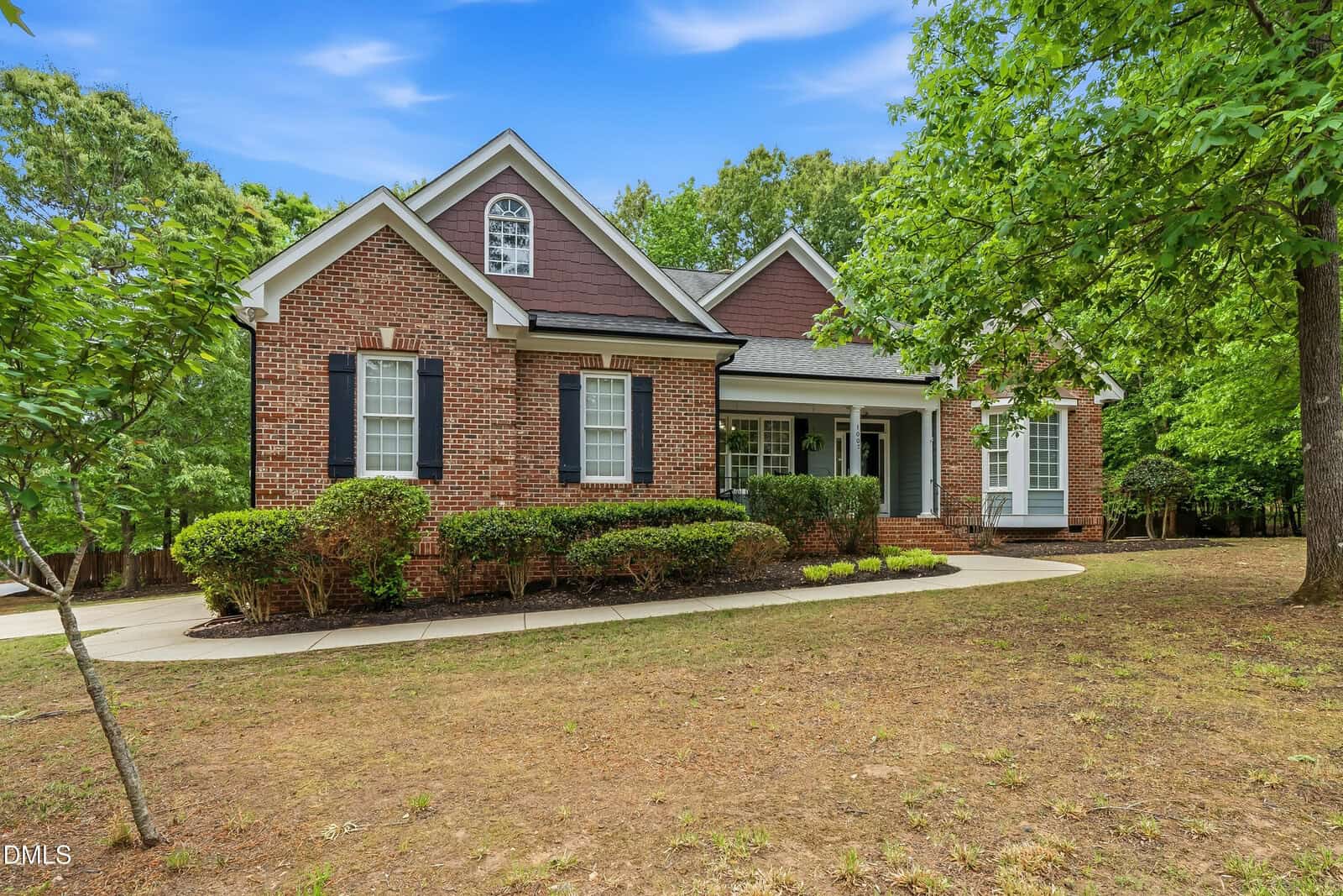 Single-story brick house with dark shutters, white trim, gabled roof, front porch, shrubs, and a curved walkway. Surrounded by trees and a patchy lawn under a bright blue sky with clouds.