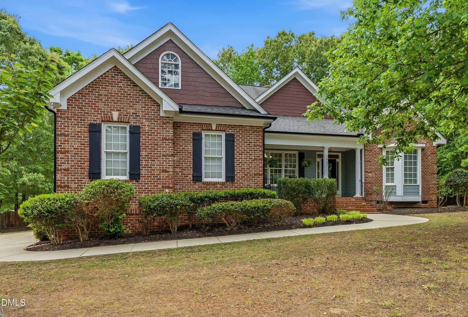 Single-story brick house with black shutters, white trim, and a front porch. Well-maintained shrubs line the walkway, and mature trees surround the home. The sky is clear and blue.