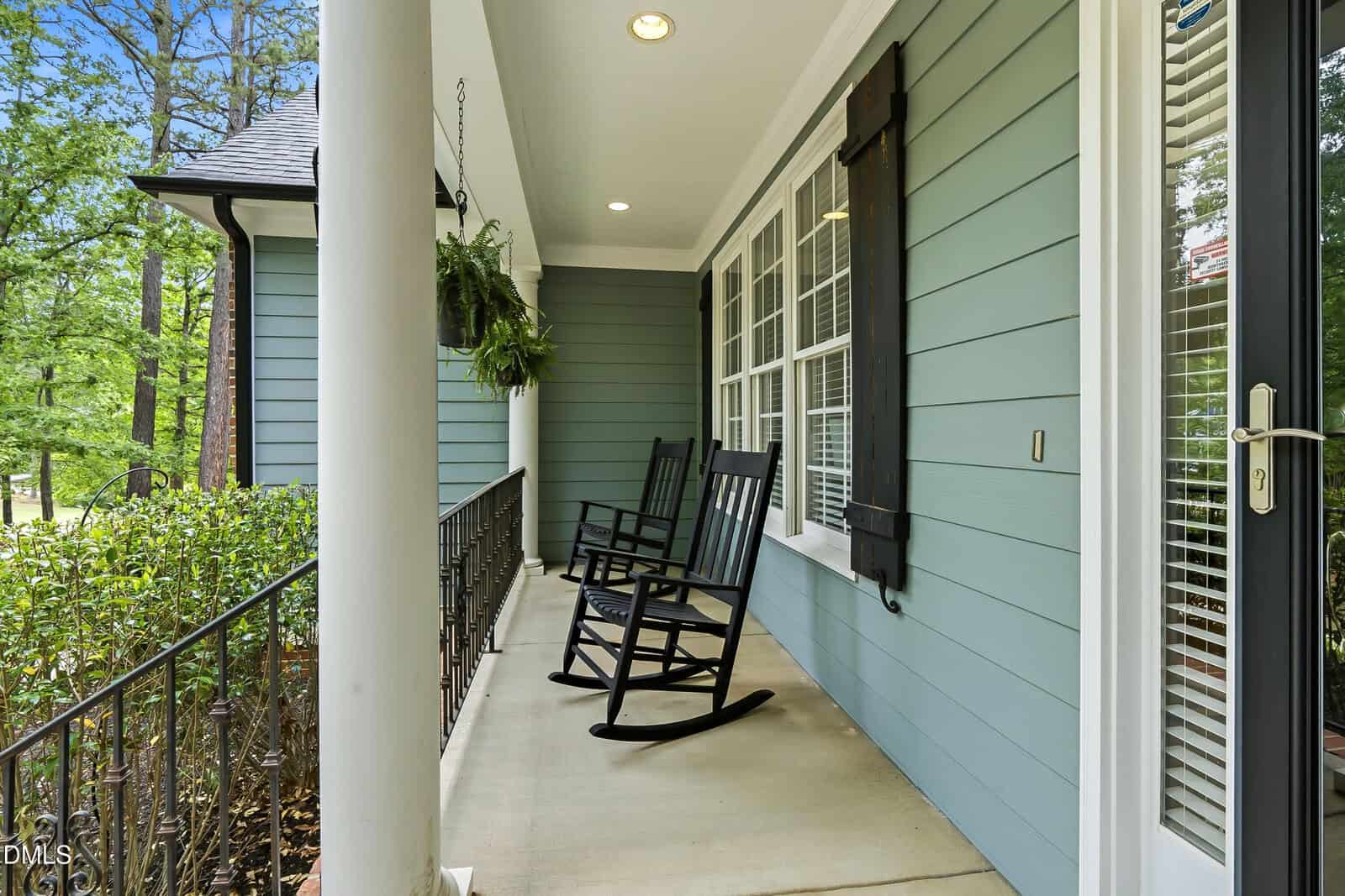 A cozy front porch with two black rocking chairs, white columns, blue siding, large windows with black shutters, a hanging plant, and greenery in the background.