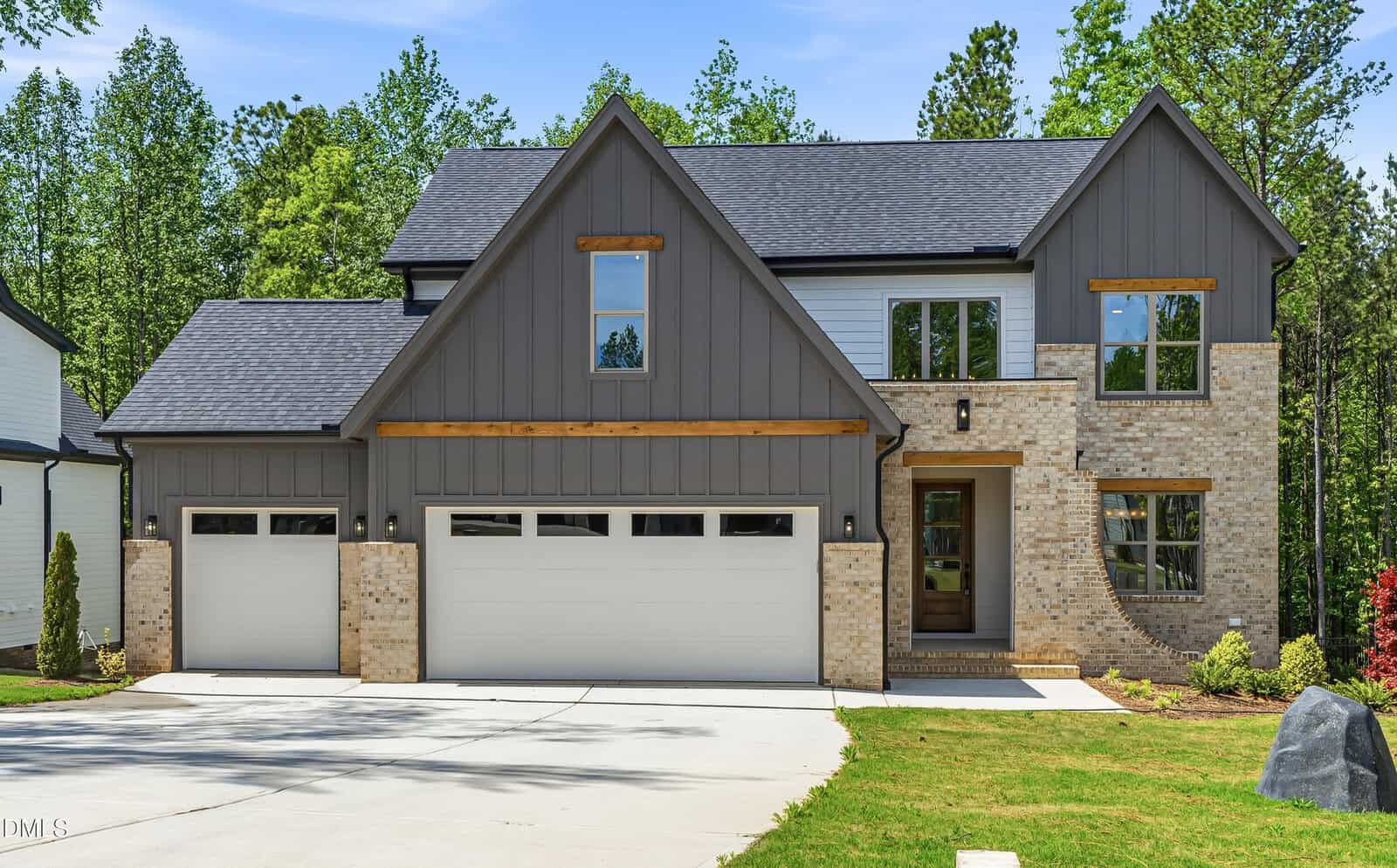 Modern two-story house with gray siding, light brick accents, and three-car garage. Large windows, gabled roof, and a covered front entry. Surrounded by trees and a neatly maintained lawn.