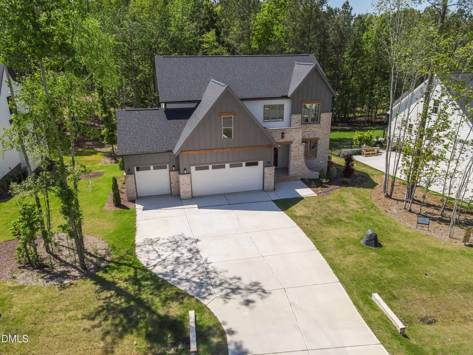 A modern two-story house with gray siding, brick accents, and a three-car garage, surrounded by trees and a well-maintained lawn with a curved driveway.