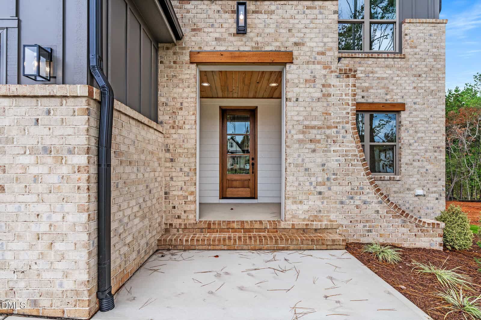 Front entrance of a modern brick house with a wooden door, covered porch, and overhanging light fixture. The sidewalk leading to the door is bordered by mulch and landscaped plants.