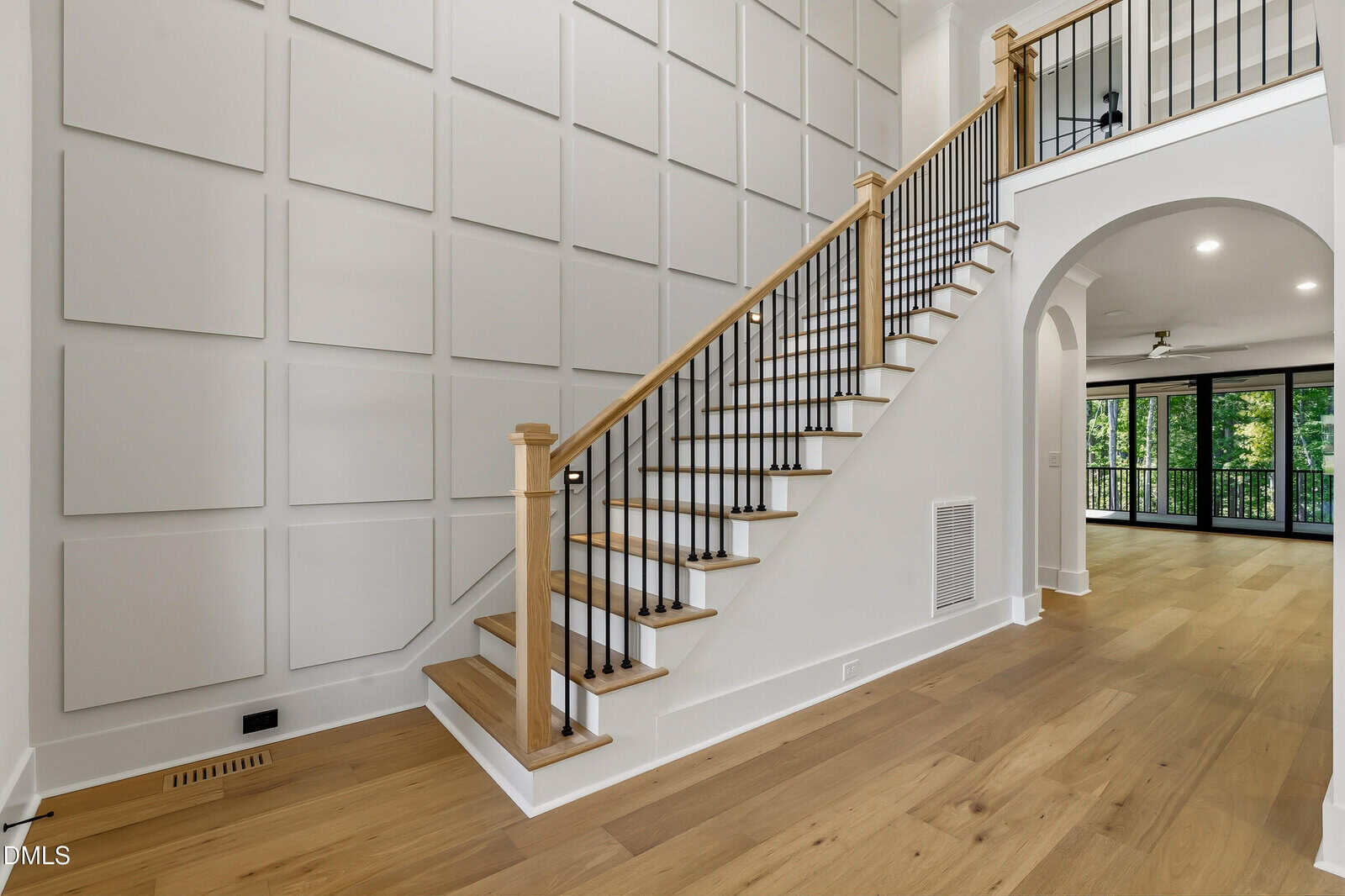 Modern interior with a wooden staircase featuring black railings, a white paneled accent wall, light wood flooring, and an arched entryway leading to a bright living area with floor-to-ceiling windows.
