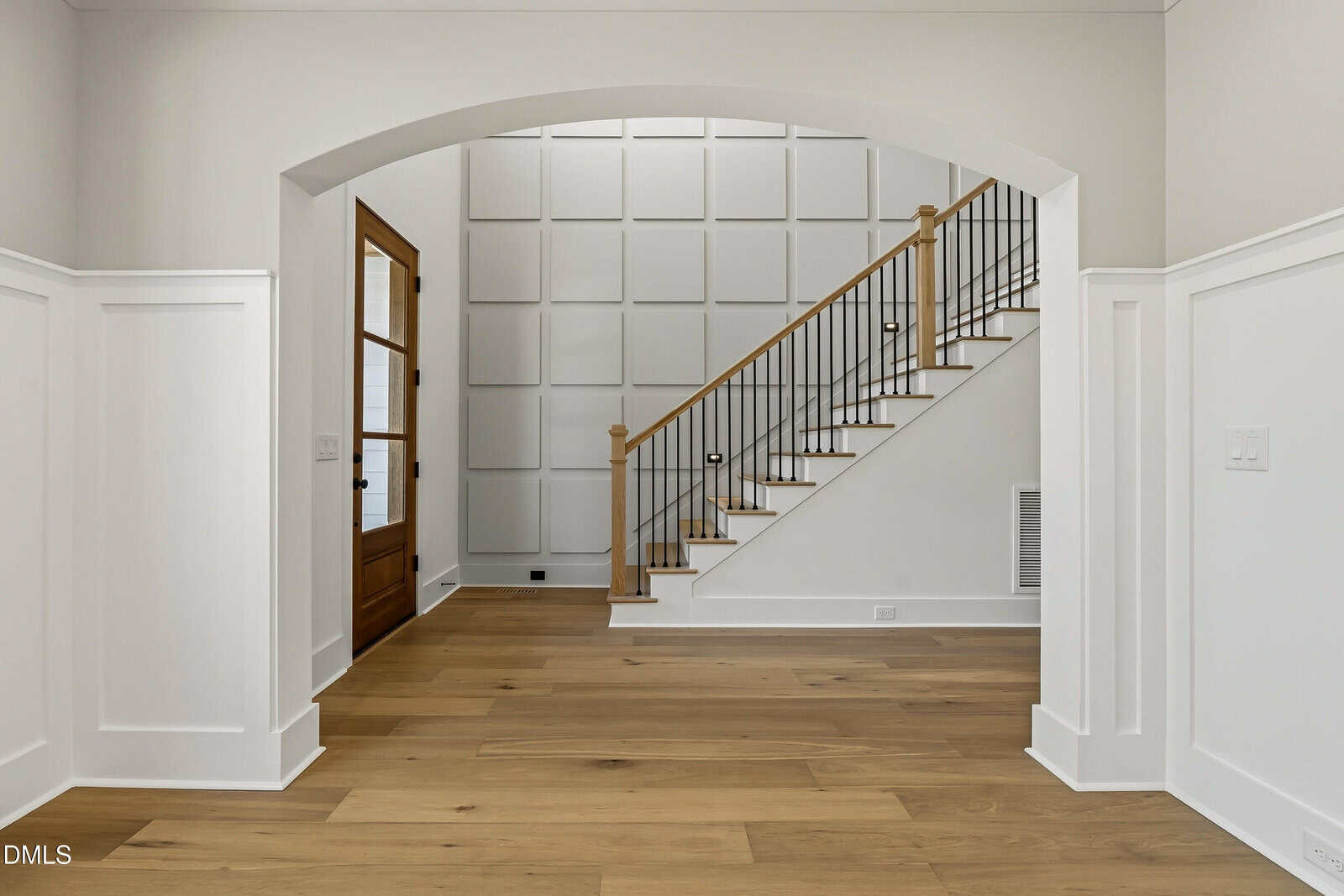 A bright entryway with light wood floors, white wainscoting, a wooden staircase with black spindles, and a large arched opening leading to a door and a paneled accent wall.