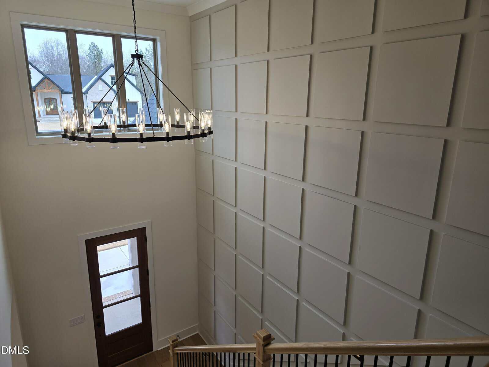 A modern entryway with a tall ceiling, grid-patterned wall paneling, a wooden stair railing, a large window, and a metal chandelier with candle-style lights.