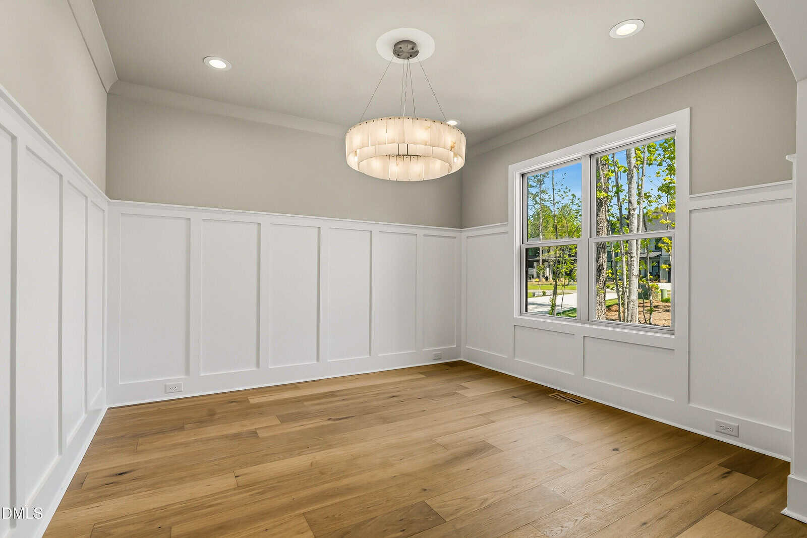 Bright empty room with light wood floors, white wainscoting, beige walls, a large window letting in natural light, and a modern circular chandelier hanging from the ceiling.