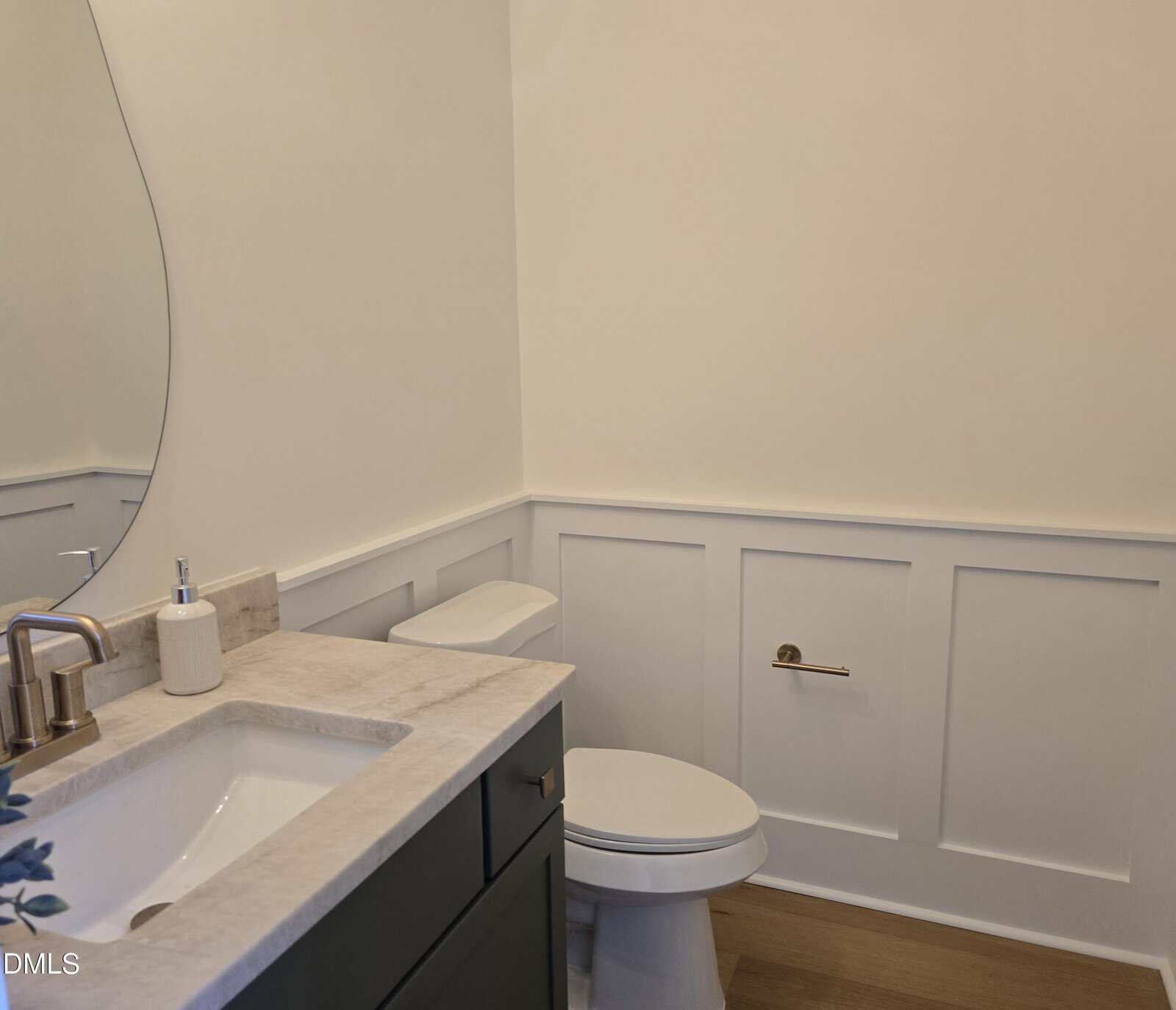 A modern bathroom with a white toilet, a sink with a soap dispenser, a curved mirror, and white paneled walls on light wood flooring.