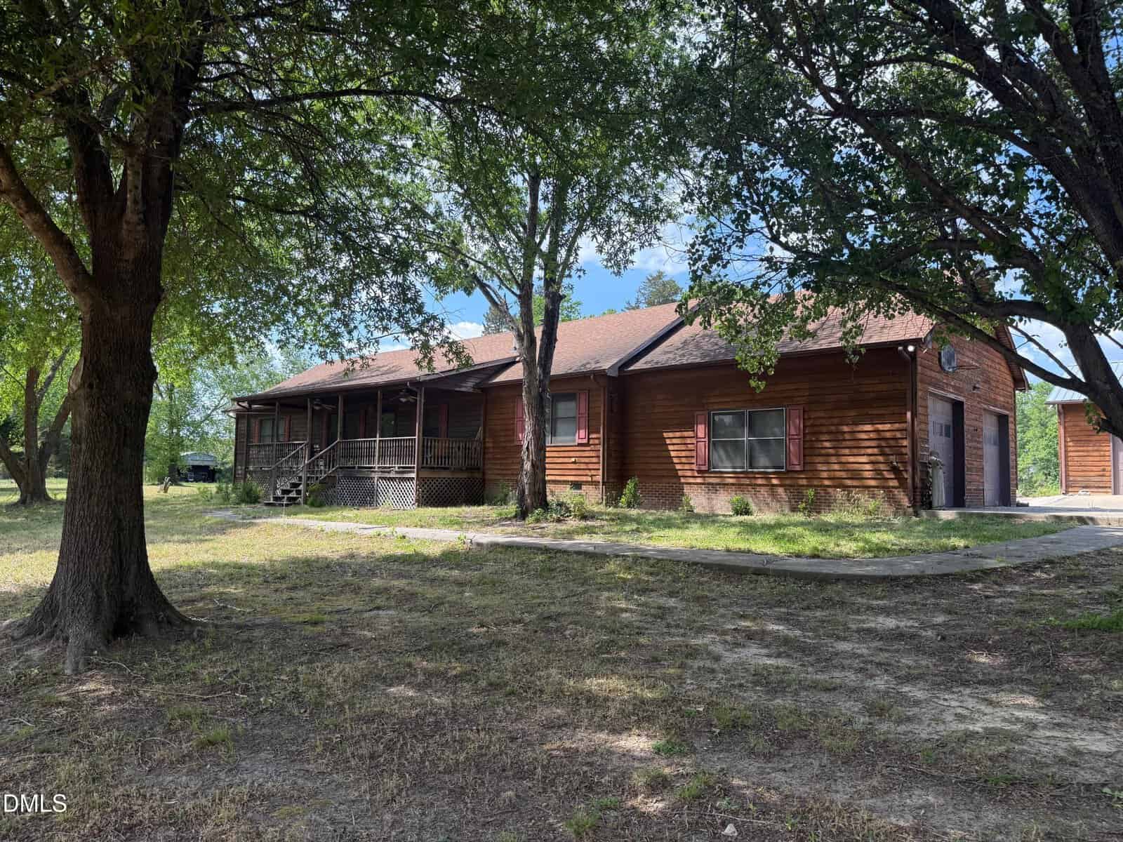 A rustic, single-story wooden house with a covered front porch and red shutters, surrounded by large trees and a patchy grass yard under a partly cloudy sky.