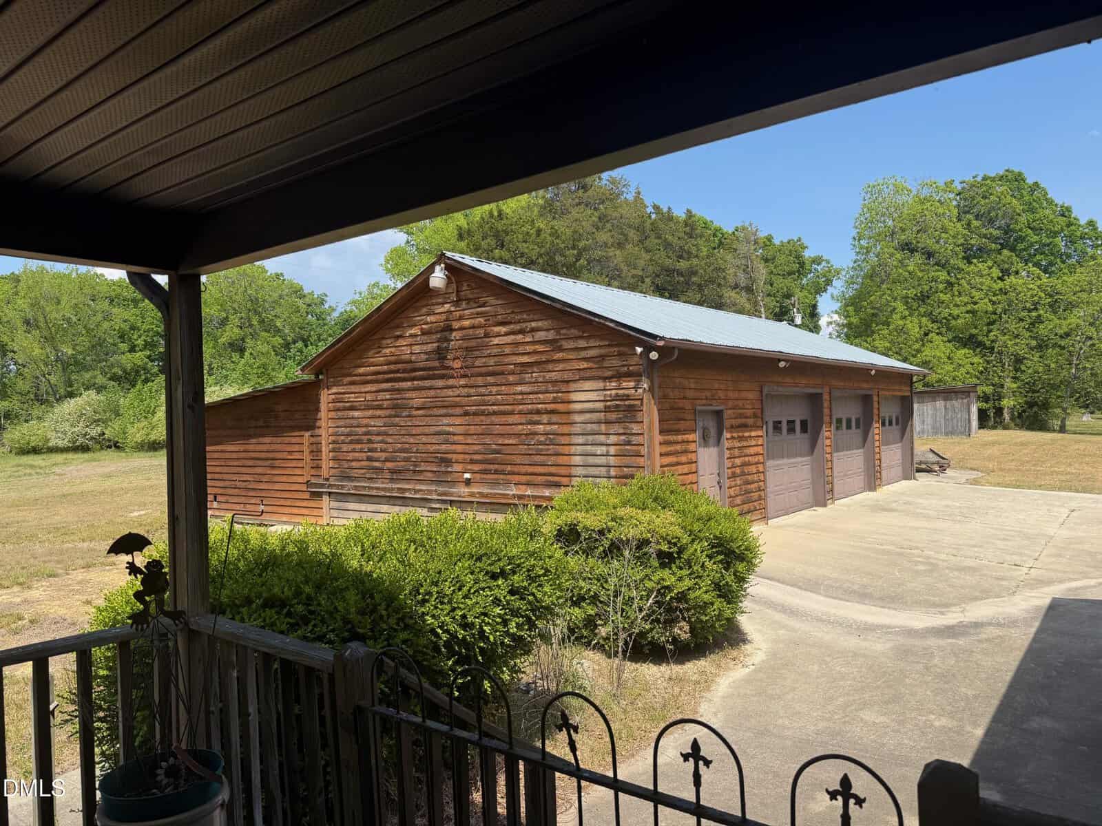 A wooden garage with three brown doors sits on a concrete driveway, surrounded by green bushes and trees, viewed from a covered porch on a sunny day.