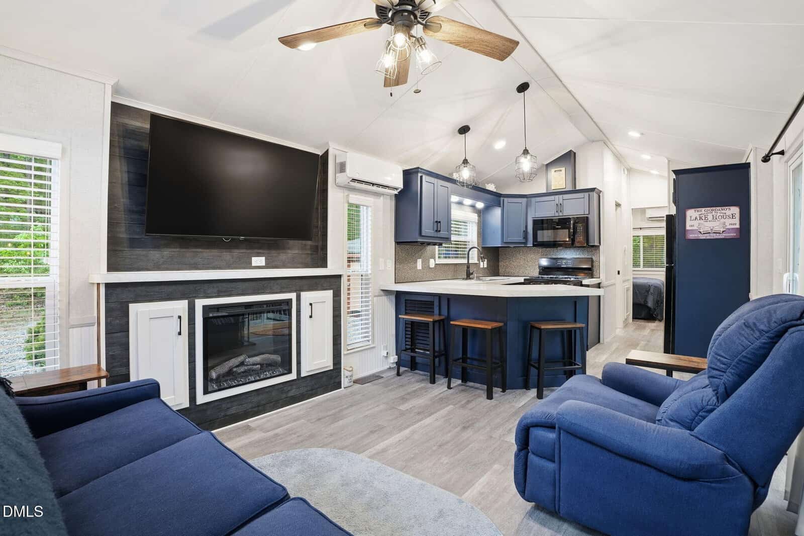 Modern tiny home interior with blue sofa and recliner, wall-mounted TV above a fireplace, kitchen with barstools, blue cabinets, stainless appliances, ceiling fan, and a hallway leading to a bedroom.