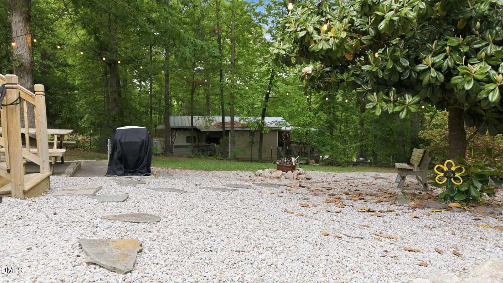 A gravel yard with a stone path, a bench, and a fire pit leads to a small cabin surrounded by tall green trees. String lights hang above, and a covered grill stands to the left near a wooden deck.