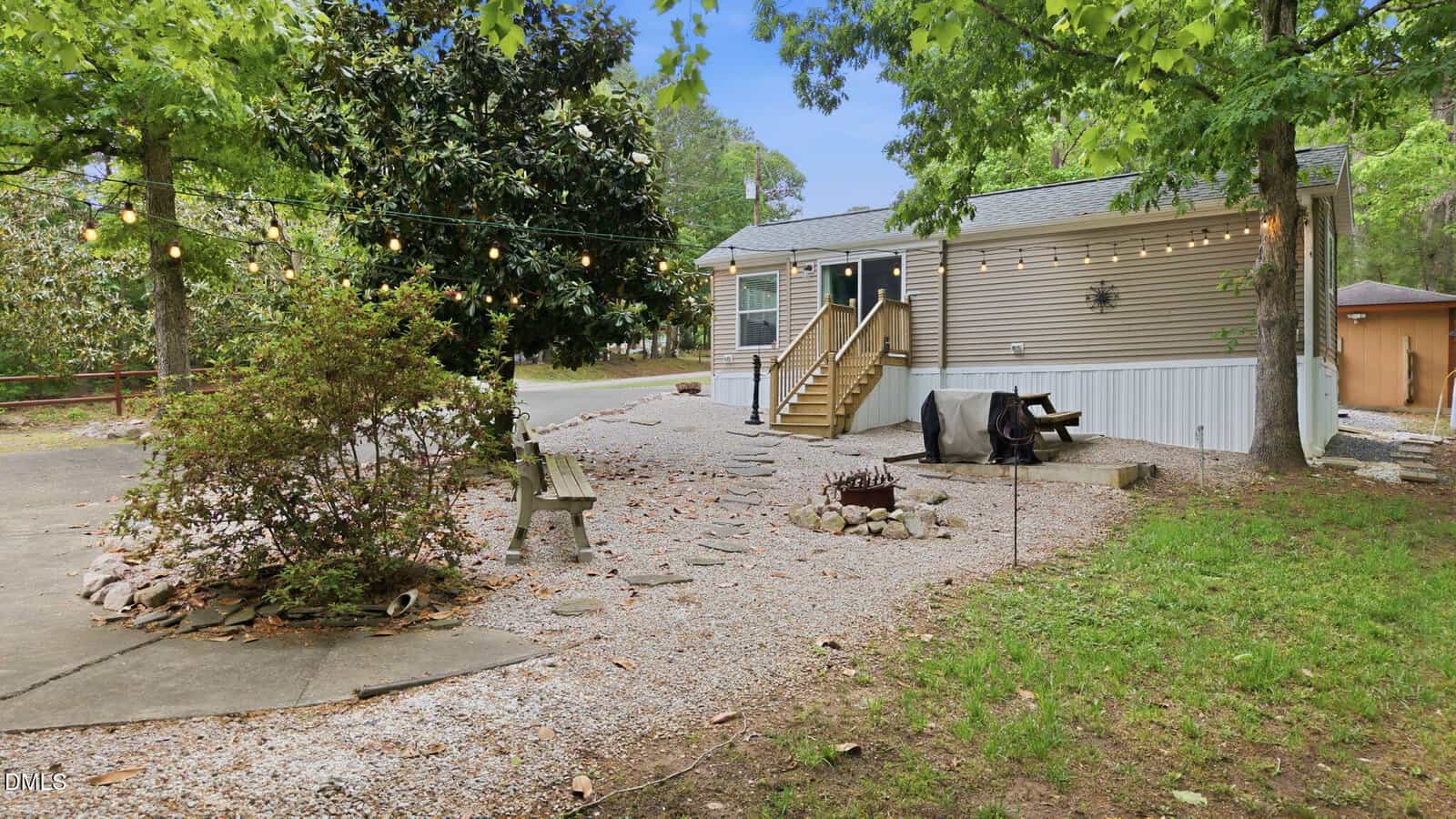 A small house with gray siding and a front porch sits among trees. The yard has string lights, a fire pit with chairs, a bench, gravel pathways, and green grass. A small shed is visible in the background.