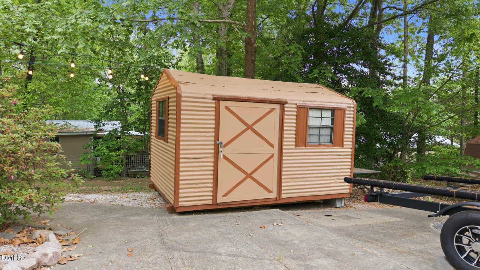 A small tan and brown corrugated metal shed with a window and door sits on a concrete pad surrounded by trees, string lights, and nearby bushes.