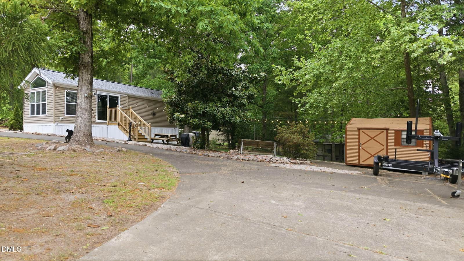 A small beige mobile home with white trim sits beside a paved driveway. There is a wooden shed, string lights, trees, and landscaping rocks in a green, wooded yard. Outdoor equipment is visible near the shed.