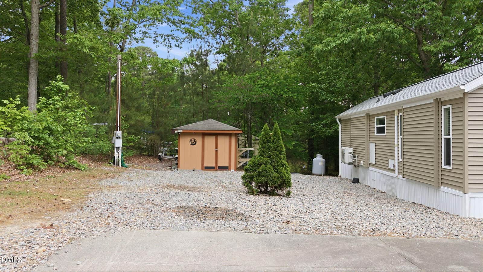 A small shed stands on a gravel driveway surrounded by trees, with the side of a tan mobile home visible on the right. A shrub sits in the center of the driveway.