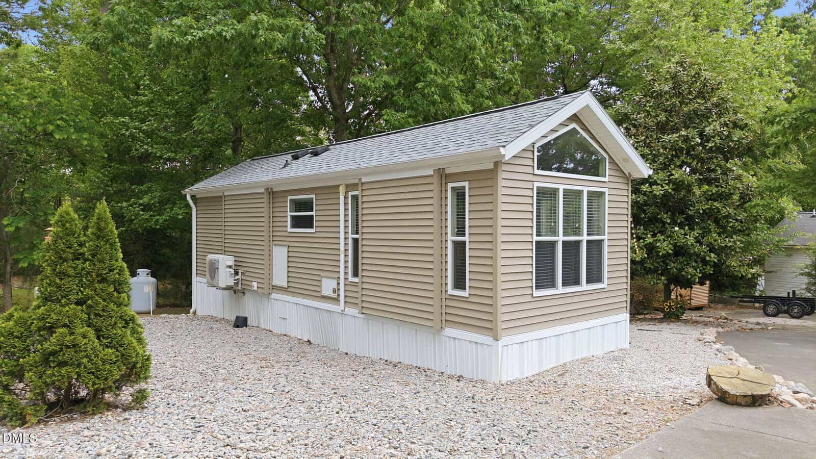 A small beige mobile home with white trim sits on a gravel lot, surrounded by trees and greenery. Large windows are at the front, and there is a driveway and trimmed bushes nearby.