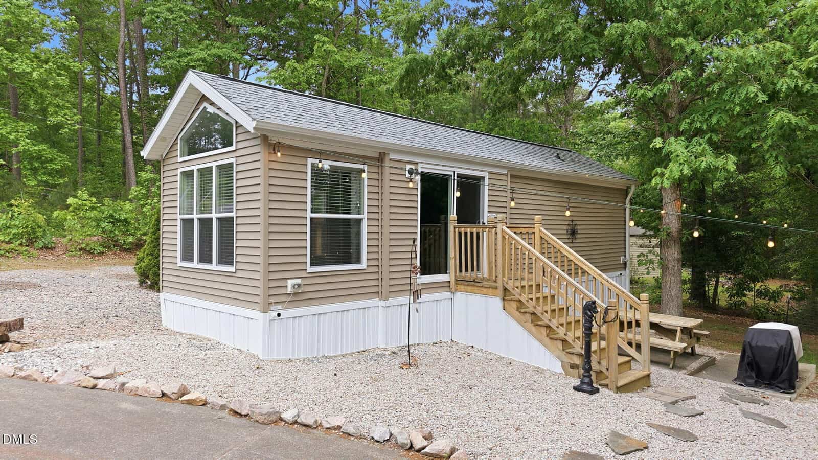 A small beige house with white trim sits on a gravel lot surrounded by trees. Wooden steps lead to the front door, and string lights hang nearby over a picnic table. The area is landscaped with rocks.
