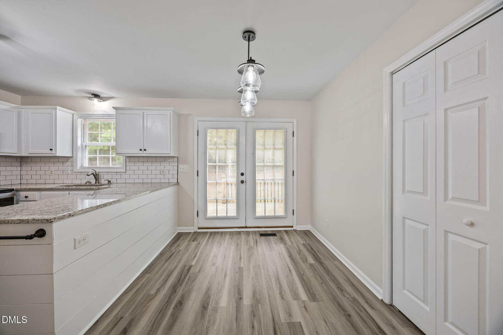 Bright kitchen with white cabinets, granite countertops, subway tile backsplash, wood flooring, and glass double doors leading outside. Modern pendant lights hang above the dining area. Closet doors are visible on the right.
