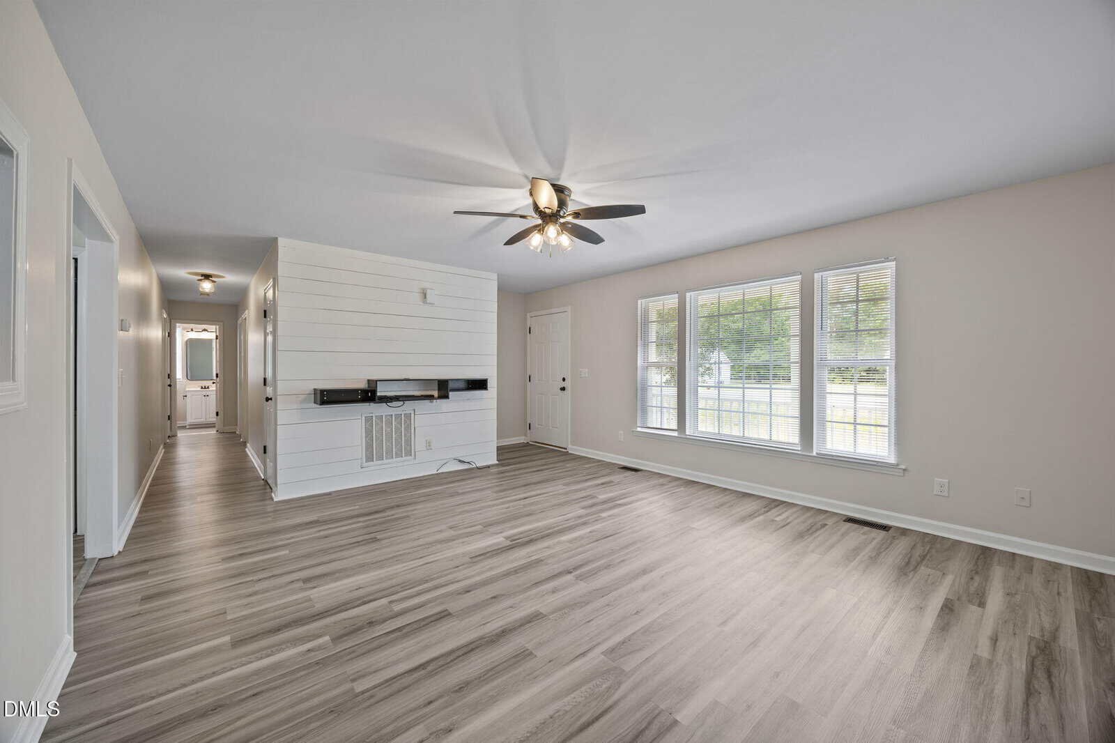 Bright living room with large windows, light wood flooring, neutral-colored walls, ceiling fan with lights, and a shiplap accent wall featuring a built-in shelf. A hallway is visible to the left.