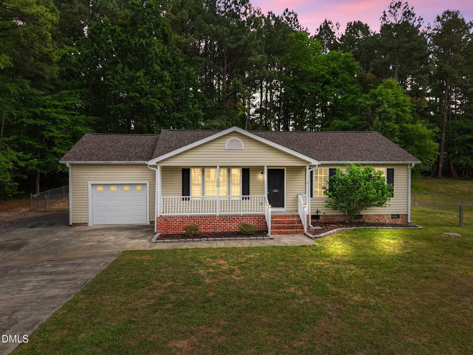 Single-story house with beige siding, a covered front porch, attached garage, and illuminated windows at dusk. Surrounded by grass lawn, mature trees, and a concrete driveway.