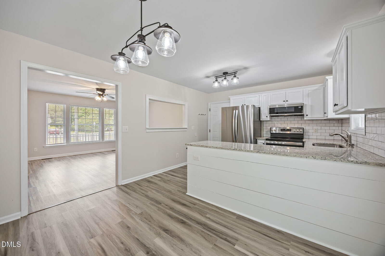 Modern kitchen with white cabinets, stainless steel appliances, subway tile backsplash, and a peninsula counter; open to a bright living room with large windows and wood-look flooring throughout.
