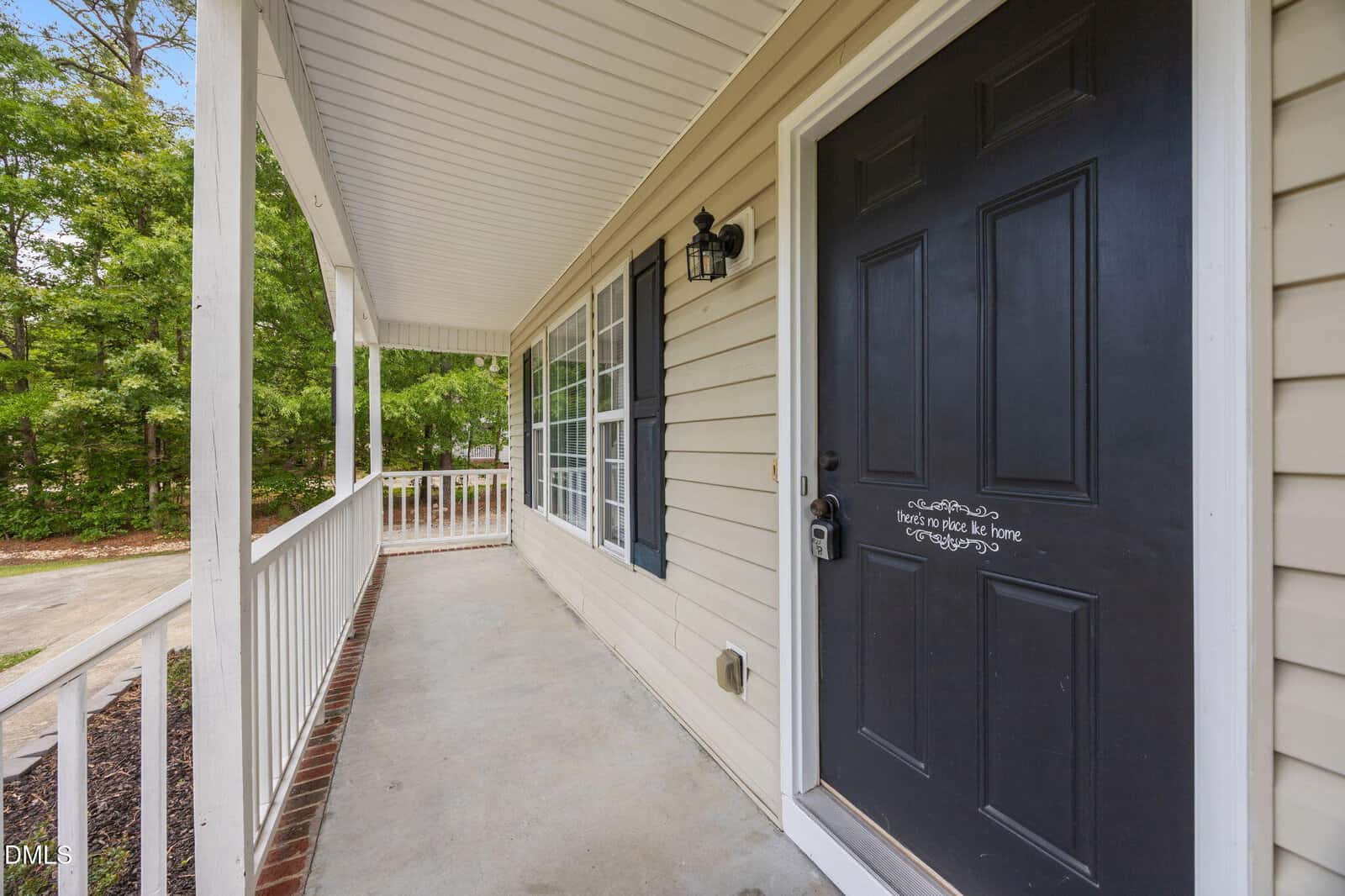 A covered front porch with white railings, beige siding, and a black front door. The door has a decorative quote, and large windows line the wall. Trees and a driveway are visible in the background.