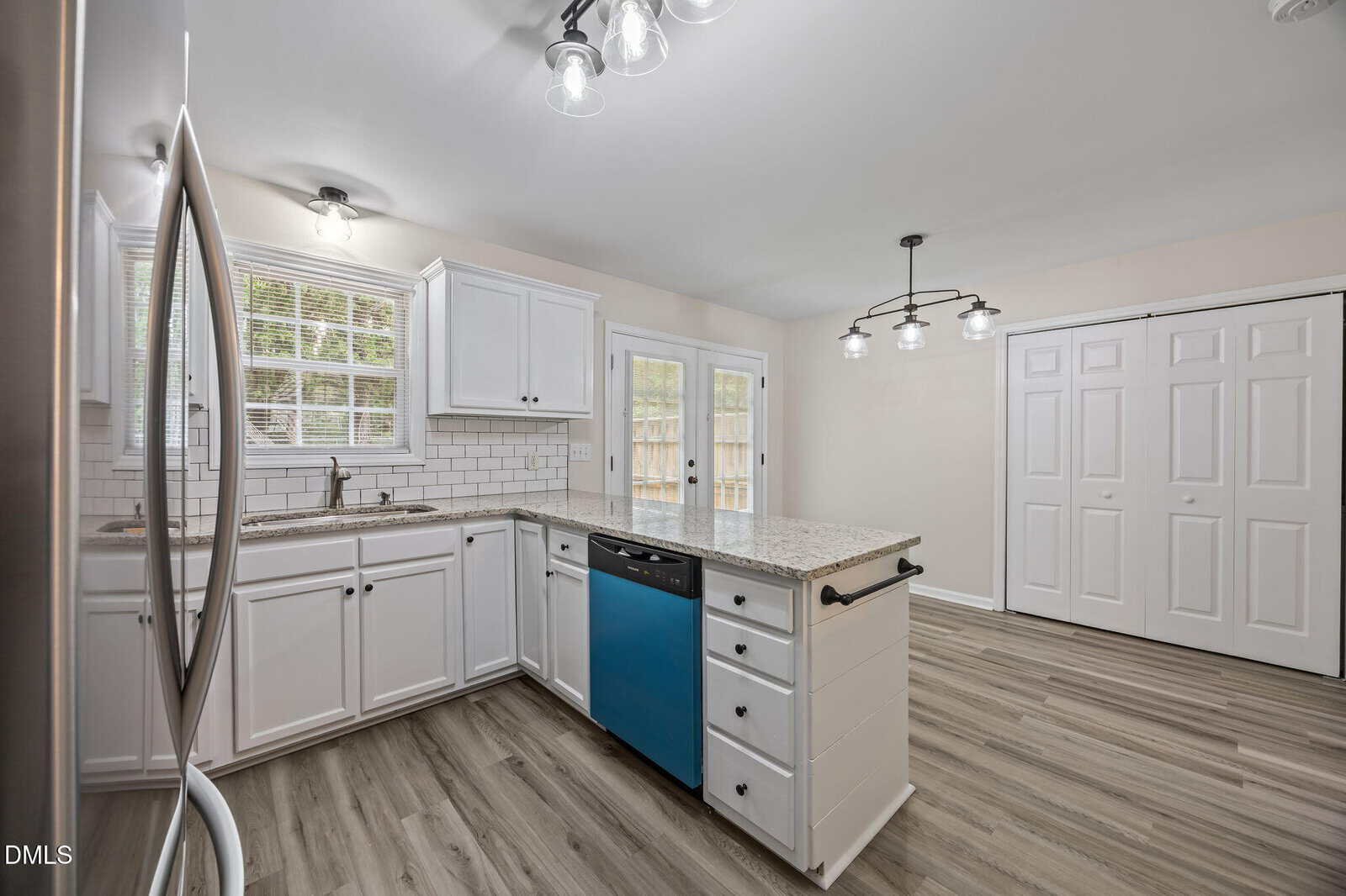 Modern kitchen with white cabinets, subway tile backsplash, granite countertops, a blue dishwasher, stainless steel fridge, light wood floors, and double white closet doors under ceiling lights. Large windows provide natural light.