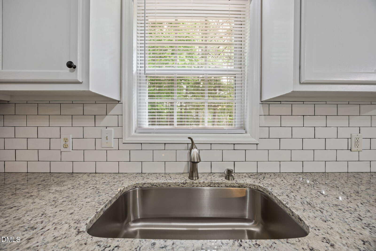 A stainless steel kitchen sink is set in a granite countertop, with white subway tile backsplash, white cabinets, and a window above with blinds partially open, showing greenery outside.