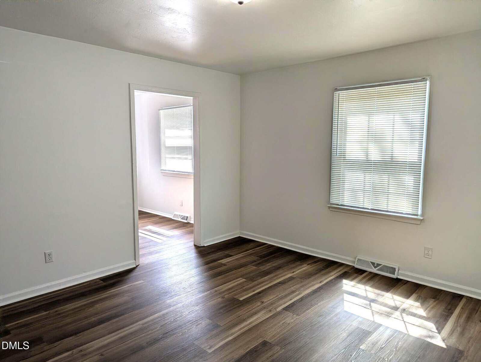 Bright, empty room with white walls, wood-style flooring, and a window with blinds, leading into an adjacent sunlit room with similar flooring and another window.