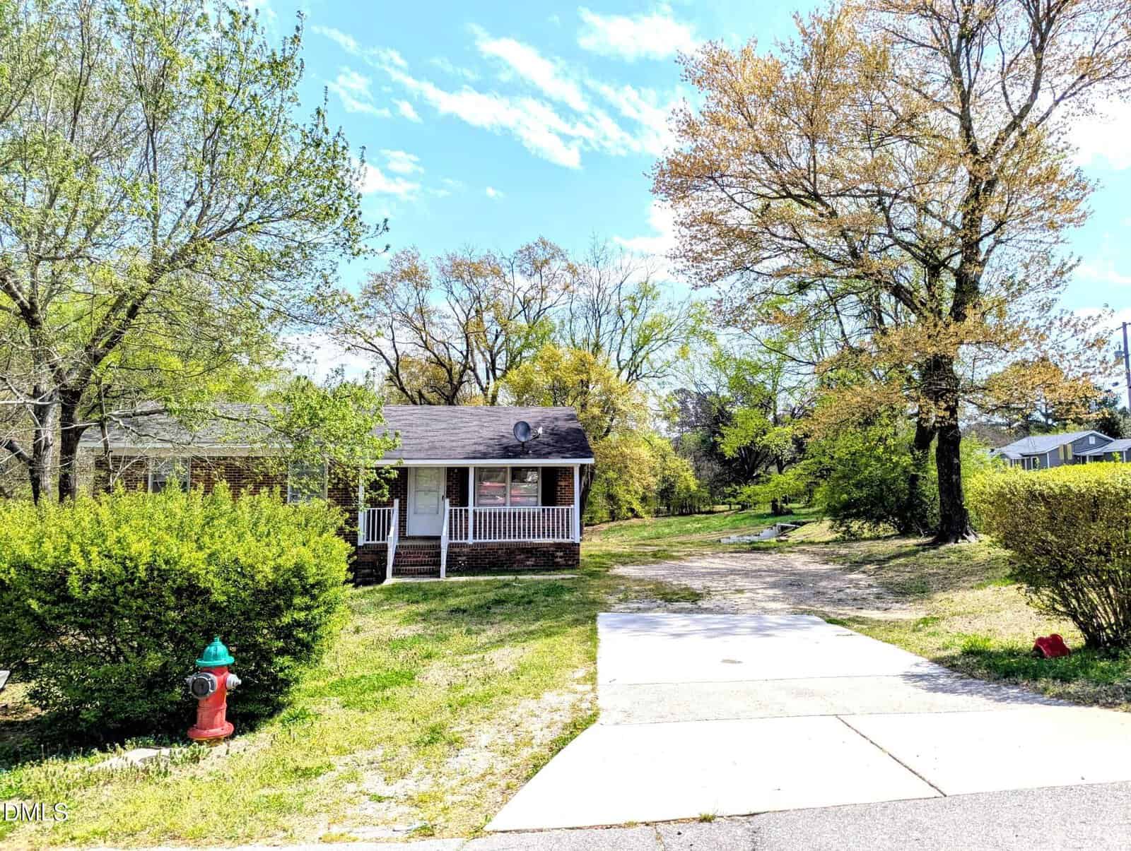 A small brick house with a covered front porch sits among tall trees and bushes. A driveway and sidewalk lead to the house, and a red fire hydrant is visible on the grass near the street. The sky is partly cloudy.